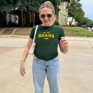 May include: A woman wearing a green t-shirt with the text "EST 1957 GHANA" and a small Ghanaian flag. She is also wearing light blue jeans and a white bag.