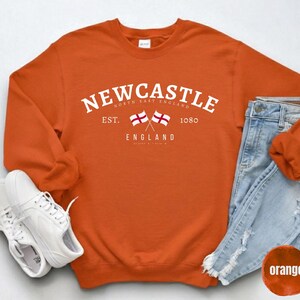 Puede incluir: Una sudadera naranja con la palabra "Newcastle" en blanco, dos banderas cruzadas y el texto "England". Se combina con vaqueros azules claros y zapatillas blancas.