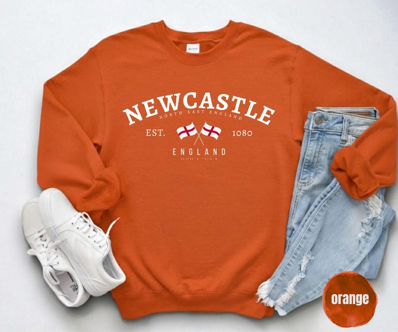 Puede incluir: Una sudadera naranja con la palabra "Newcastle" en blanco, dos banderas cruzadas y el texto "England". Se combina con vaqueros azules claros y zapatillas blancas.