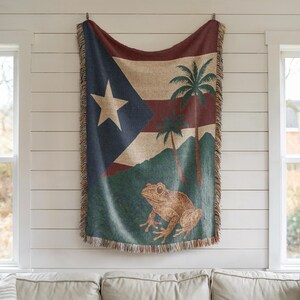 May include: A woven throw blanket featuring the flag of Puerto Rico, with a large white star on a blue triangle, red stripes, and palm trees. A brown frog is at the bottom. The blanket has a fringed edge and is hanging on a white wall.