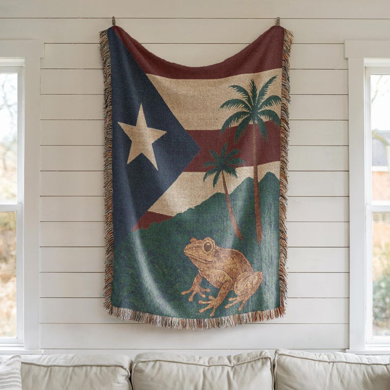 May include: A woven throw blanket featuring the flag of Puerto Rico, with a large white star on a blue triangle, red stripes, and palm trees. A brown frog is at the bottom. The blanket has a fringed edge and is hanging on a white wall.