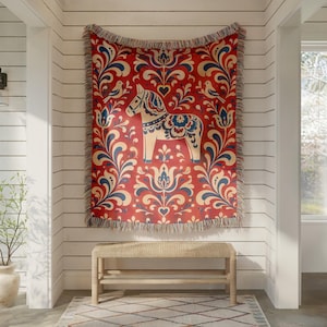 May include: A woven throw blanket featuring a red background with a repeating pattern of blue and cream floral motifs and birds. A central cream-colored Dala horse figure is decorated with blue and red floral details. The edges have a fringed trim.