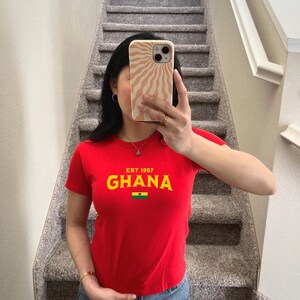 May include: Red t-shirt with a yellow graphic that says "EST 1957 GHANA" and a small Ghanaian flag.