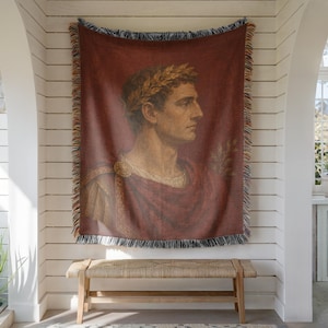 May include: A rectangular woven throw blanket featuring a profile portrait of a man wearing a laurel wreath and a deep red toga. The blanket has a fringed edge with multicolored threads and displays a classical artistic style in shades of red.