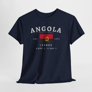 May include: A navy blue t-shirt with a white graphic design that says "ANGOLA" with the text "REPUBLICA DE ANGOLA" underneath. The graphic also includes the Angolan flag, the text "EST. 1975", and the text "LUANDA 8.8390° S, 13.2894° E".