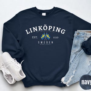 May include: Navy blue sweatshirt with the text "LINKÖPING" and "SWEDEN" in white. Features two Swedish flags and the text "EST. 1120". Includes a pair of white sneakers and light blue jeans.
