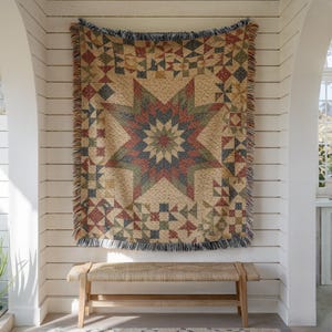 May include: A woven throw blanket featuring a large central star pattern in muted shades of red, blue, and green on a cream background. The blanket has a decorative fringe along the edges and is displayed hanging vertically against a white wall.