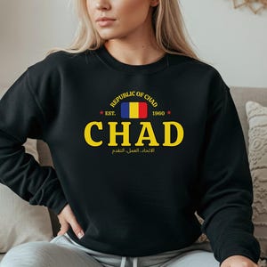 May include: Black sweatshirt with a yellow and white graphic that says "CHAD" with the flag of Chad. The text "REPUBLIC OF CHAD" and "EST. 1960" are above the word "CHAD". Below the word "CHAD" is text in Arabic.