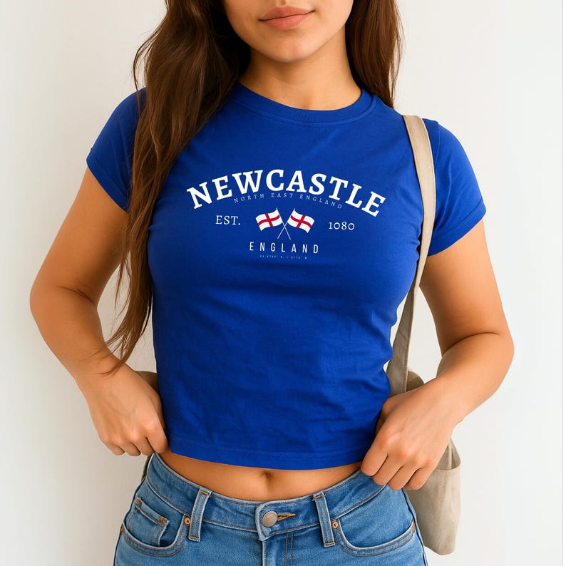 May include: Royal blue crop top with the word "Newcastle" in white arched text. Below the text are two crossed flags and the word "England". The text "North East England" and "Est. 1080" are also printed on the shirt.