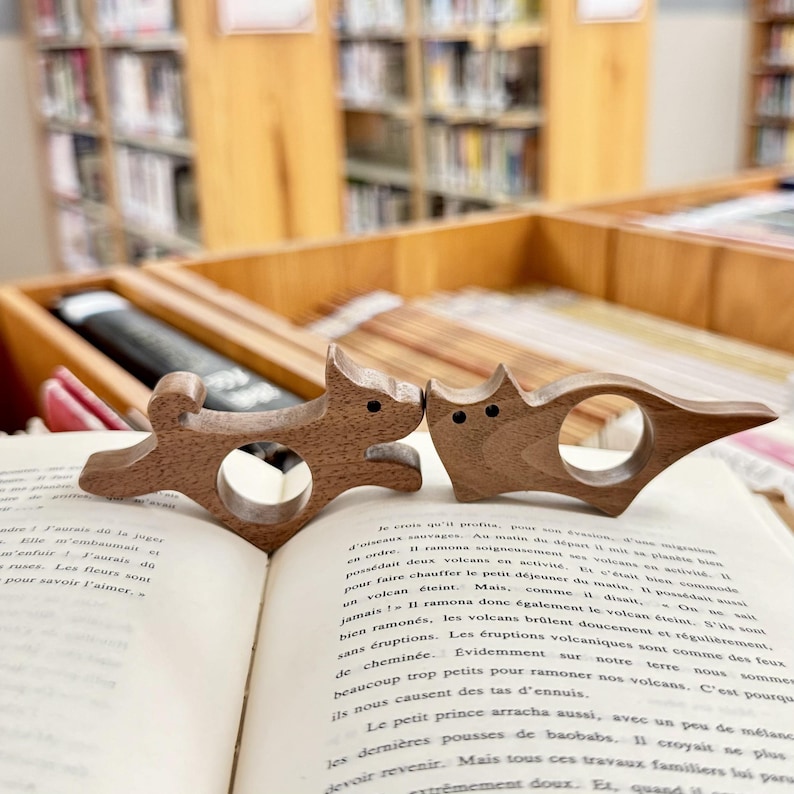 May include: Two wooden animal ring holders in the shape of a cat and a dog. The cat is brown and has a white face. The dog is brown and has a white face. The ring holders are sitting on a book.