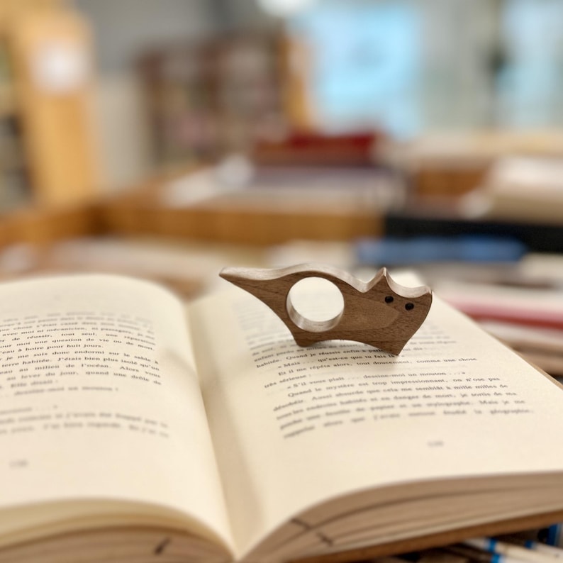 May include: A wooden cat-shaped bookmark with a ring for holding a page. The bookmark is sitting on an open book.