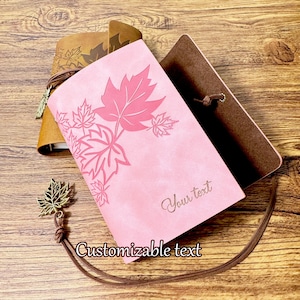 May include: A pink leather journal with a brown leather cover and a brown leather strap. The journal has a maple leaf design and the text "Your text" is printed on the front cover. The journal comes with a brown leather strap and a gold-colored maple leaf charm.
