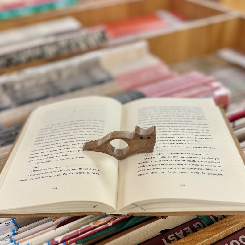 May include: A wooden cat-shaped bookmark with a ring to hold a page. The bookmark is resting on an open book.