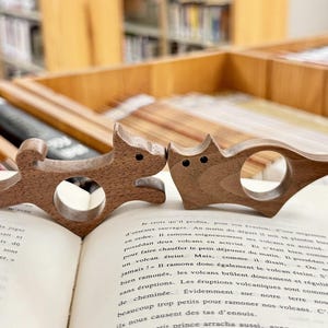 May include: Two wooden bookends shaped like animals. The bookends are brown and have a natural wood finish. The bookends are sitting on an open book.