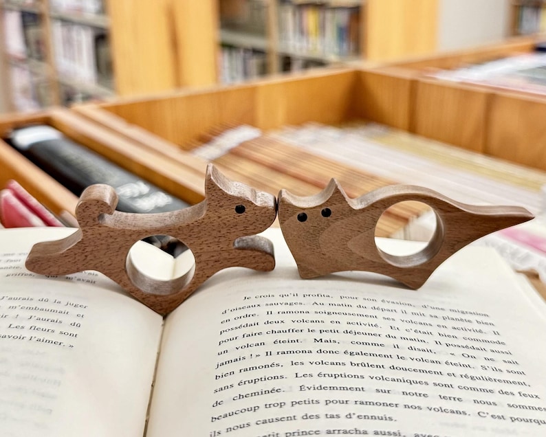 May include: Two wooden bookends shaped like animals. The bookends are brown and have a natural wood finish. The bookends are sitting on an open book.