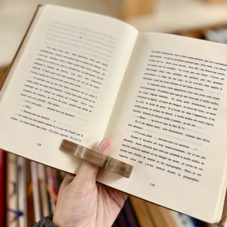 May include: A close-up of an open book with a wooden page holder. The book is open to a page with text about a man who is lost and a drawing of a sheep. The text reads "S'il vous pla&icirc;t... dessine-moi un mouton..."
