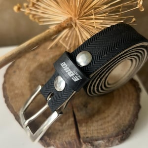 Peut inclure: Ceinture noire avec une boucle argentée et deux rivets argentés. La ceinture présente un motif texturé et le texte "E-BIKE READY" est visible. La ceinture est enroulée sur une surface en bois.
