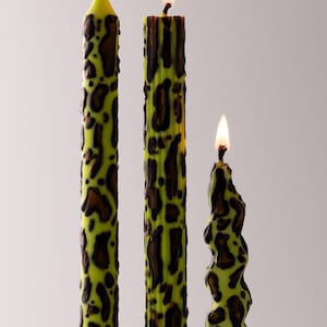 May include: Three lit candles with a green and brown leopard print design. The candles are different shapes, including a straight, a ribbed, and a twisted design. The flames are yellow and orange.