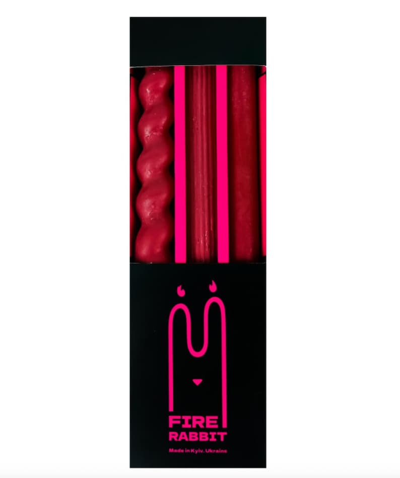 May include: A black box containing red candles of various shapes. The box features the text "FIRE RABBIT" in pink, along with a rabbit logo. The candles are arranged vertically, with a twisted candle on the left.