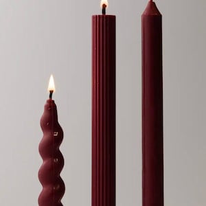 May include: Three burgundy candles with lit flames. The candles vary in design: a spiral, a ribbed column, and a smooth, tapered pillar. The candles are set against a plain white background.