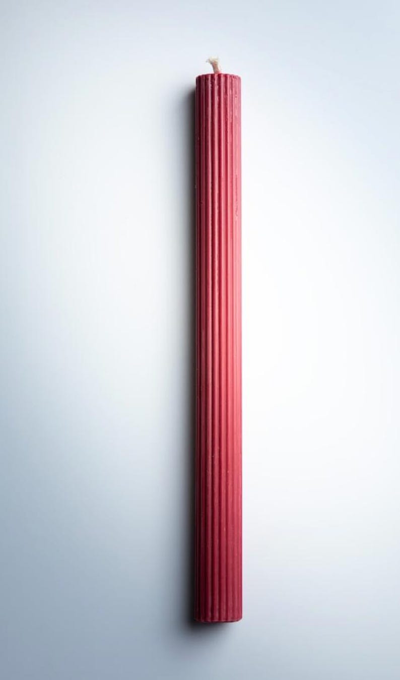 May include: A tall, red pillar candle with a ribbed design. The candle has a simple, elegant form and a wick at the top. The candle is a deep red color and is set against a white background.
