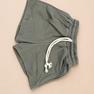 May include: A pair of sage green shorts with an elastic waistband and a white drawstring. The shorts are made of a soft, casual fabric, perfect for lounging or light activity. The shorts are folded neatly.