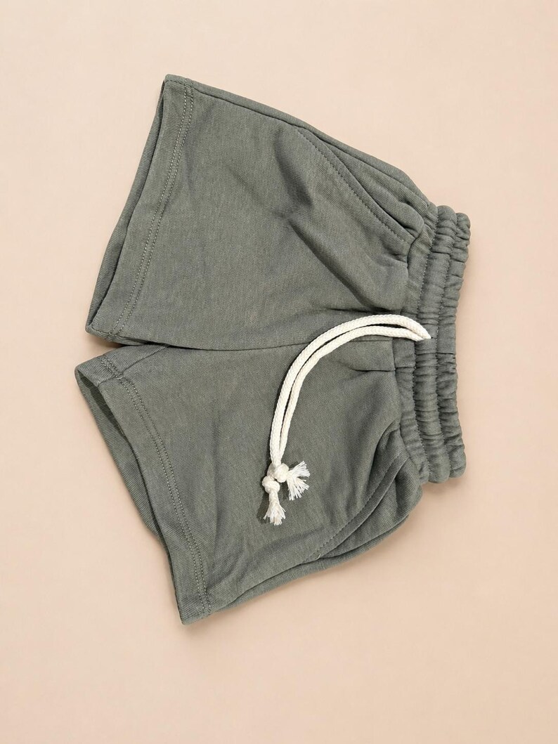 May include: A pair of sage green shorts with an elastic waistband and a white drawstring. The shorts are made of a soft, casual fabric, perfect for lounging or light activity. The shorts are folded neatly.