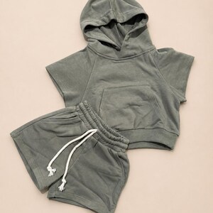 May include: A sage green hooded sweatshirt and matching shorts set. The short-sleeved hoodie has a front pocket and a drawstring hood. The shorts have an elastic waistband with a white drawstring. The set is made of a soft, comfortable material.