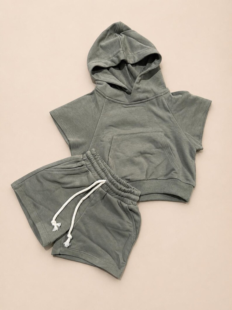 May include: A sage green hooded sweatshirt and matching shorts set. The short-sleeved hoodie has a front pocket and a drawstring hood. The shorts have an elastic waistband with a white drawstring. The set is made of a soft, comfortable material.