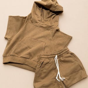 May include: A two-piece set of children's clothing in a muted brown color. The set includes a hooded, short-sleeved top with a front pocket and matching shorts with a drawstring waist. The fabric appears to be soft and comfortable.