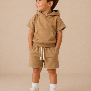 May include: A child wearing a tan short-sleeved hoodie and matching shorts with a drawstring. The outfit includes white sneakers and socks. The child is smiling and looking to the side.