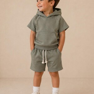 May include: A young child wearing a sage green short-sleeved hoodie and matching shorts. The shorts have a drawstring waist. The child is also wearing white socks and white sneakers with velcro straps. The background is a solid, neutral color.