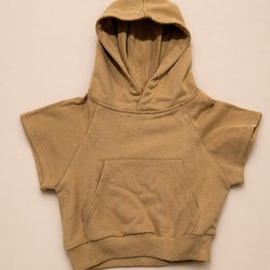 May include: A tan-colored hooded sweatshirt with short sleeves. The sweatshirt features a front pocket and a ribbed hem. The hood is lined and the garment is made of a soft, comfortable material. This casual top is suitable for children.