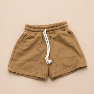 May include: Tan-colored shorts with an elastic waistband and a white drawstring. The shorts have two side pockets and are made of a soft, comfortable material. These casual shorts are perfect for everyday wear.