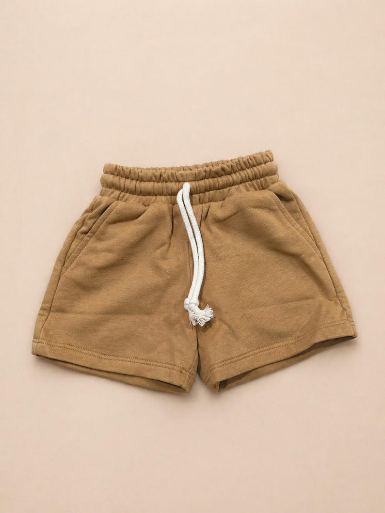 May include: Tan-colored shorts with an elastic waistband and a white drawstring. The shorts have two side pockets and are made of a soft, comfortable material. These casual shorts are perfect for everyday wear.