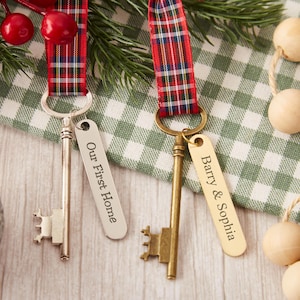 May include: Two decorative skeleton keys with engraved tags. One tag reads "Our First Home" and the other reads "Barry & Sophia." The keys are attached to red and blue plaid ribbons, with greenery and red berries in the background.