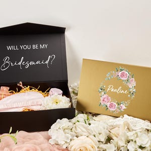 Puede incluir: Se muestran dos cajas de regalo, una negra y otra dorada. La caja negra está abierta, revelando una máscara para dormir y otros artículos. El texto "WILL YOU BE MY Bridesmaid?" está en la tapa. La caja dorada tiene un diseño floral y el nombre "Pauline".