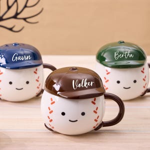 Può includere: Tre tazze a tema baseball con coperchi, ognuna con un volto sorridente e cuciture da baseball. I coperchi sono blu, marroni e verdi, con i nomi "Gavin", "Volker" e "Bertha" in scrittura bianca. Le tazze hanno manici marroni.