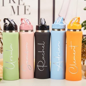 Puede incluir: Cinco botellas de agua coloridas con nombres: Ursula, Samson, Rachel, Adrian y Clement. Cada botella tiene un color diferente: verde, rosa, negro, azul y naranja. Los nombres están en plata. Las tapas son negras.