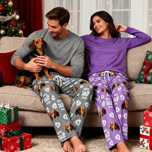 Puede incluir: Una pareja con pantalones de pijama a juego con caras de perro salchicha. El hombre lleva una camiseta gris de manga larga y sostiene un perro salchicha. La mujer lleva una camiseta morada de manga larga. Regalos de Navidad y un árbol decorado.