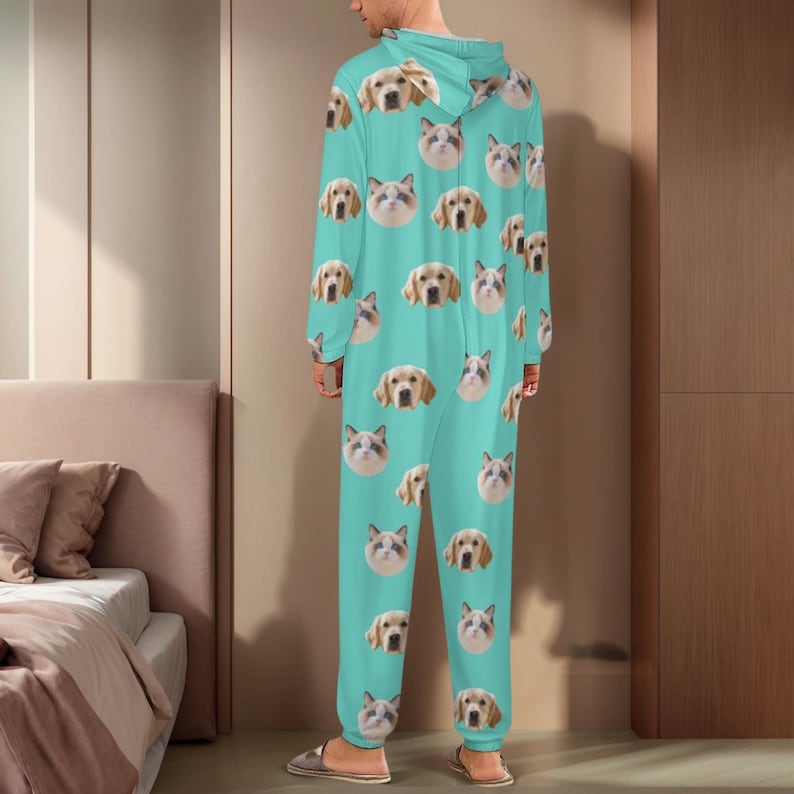May include: A turquoise hooded jumpsuit featuring a repeating pattern of dog and cat faces. The one-piece garment has long sleeves and a relaxed fit. The person is wearing gray and brown slip-on shoes.