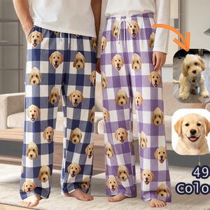 May include: Two pairs of pyjama bottoms featuring a repeating pattern of a golden puppy face. One pair has a blue and white buffalo check pattern, and the other has a purple and white buffalo check pattern. Both pairs include a white drawstring waist.