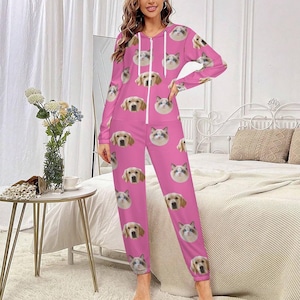 May include: A pink jumpsuit featuring a repeating pattern of dog and cat faces. The jumpsuit has a hooded top with a white zipper and long sleeves. The model is standing in a bedroom setting.