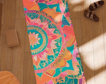 May include: A colorful yoga mat with a floral mandala design in shades of pink, orange, teal, and gold. The mat is rectangular and lies on a wooden floor. The Aura Yoga logo is visible in the bottom right corner.