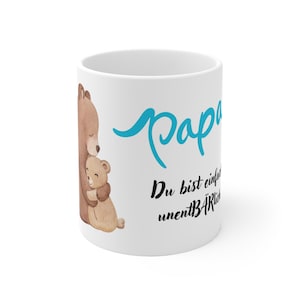 Könnte beinhalten: Weißer Keramikbecher mit einer Illustration eines Bären, der einen kleineren Bären umarmt. Der Becher trägt das Wort "Papa" in türkisfarbener Schrift, darunter deutscher Text. Ein herzerwärmendes Geschenk.
