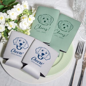 May include: Four beverage insulators in shades of gray and green, each featuring a dog illustration and the word "Cheers!" in script. The text "Love, Bodhi" and "Love, Ravioli" are also printed on the insulators. The insulators are on a white plate.