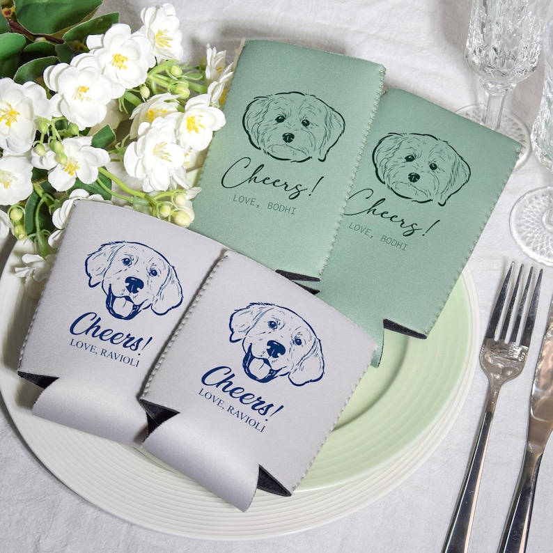 May include: Four beverage insulators in shades of gray and green, each featuring a dog illustration and the word "Cheers!" in script. The text "Love, Bodhi" and "Love, Ravioli" are also printed on the insulators. The insulators are on a white plate.