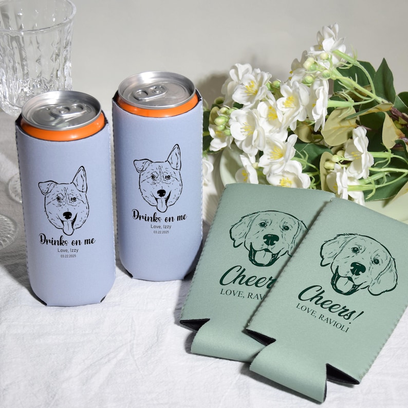 May include: Two light blue can coolers with dog illustrations and the text "Drinks on me". Two green can coolers with dog illustrations and the text "Cheer" and "Cheers!". White flowers and a glass are in the background.