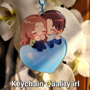 May include: A heart-shaped keychain featuring a cartoon couple embracing. The keychain has a blue heart base and a blue chain. The text "Keychain Quality irl" is at the bottom of the image. The couple has brown hair and is wearing blue and white clothing.
