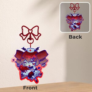 May include: A red and white anime-style nurse charm with a red bow and a lobster clasp. The charm features a cartoon character with red hair, a nurse's cap, and a syringe. The back of the charm is also visible.